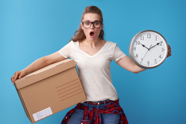 shocked young woman with cardboard box showing clock on blue