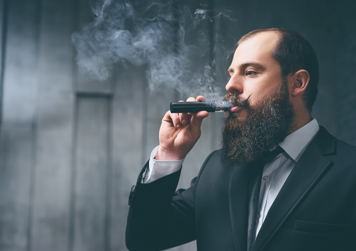 Elegant vaper. Handsome young bearded man in suit smoking vaporizer electronic cigarette.
