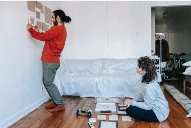 A man arranging cut cardboard pieces onto the wall to help him arrange his artwork while a woman is sitting on the floor watching him.