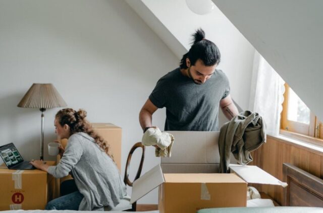A man decluttering and packing items in big cardboard boxes while the woman is using her laptop in the background